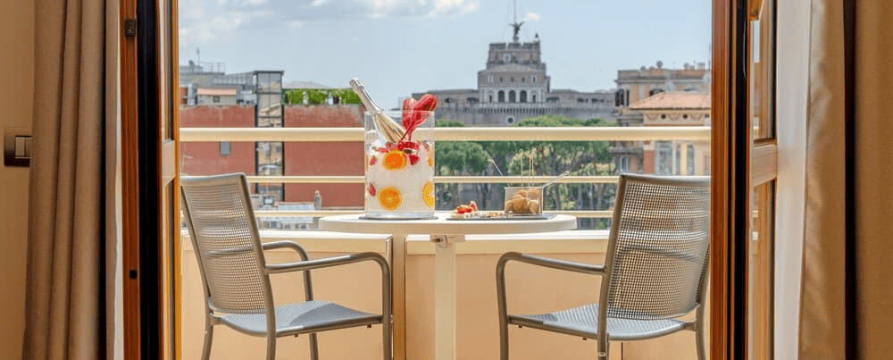 Balcony with two chairs and a table featuring drinks, overlooking a city skyline with historic build.