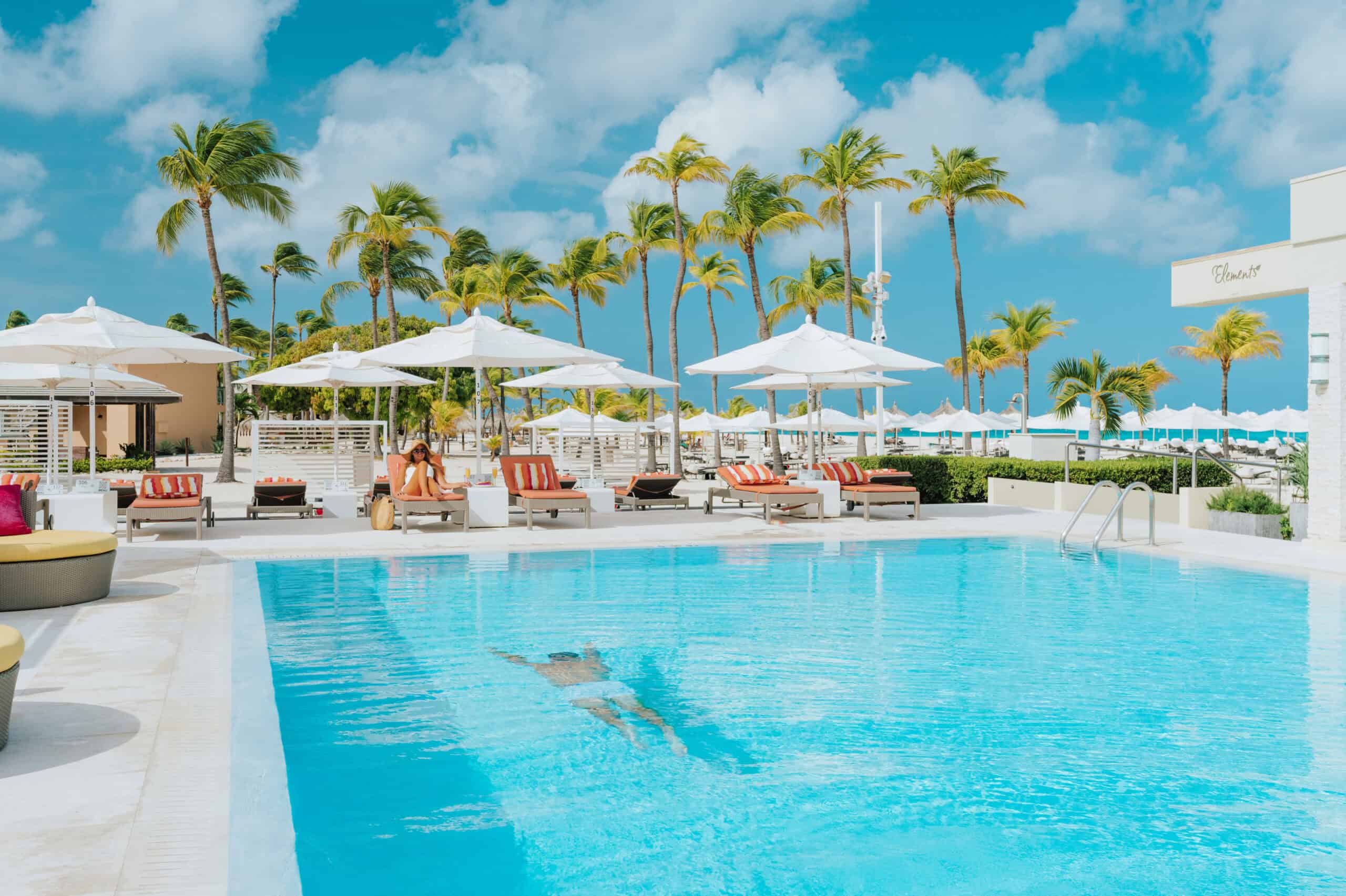 Relaxing tropical resort pool with palm trees, lounge chairs, and umbrellas under a blue sky.