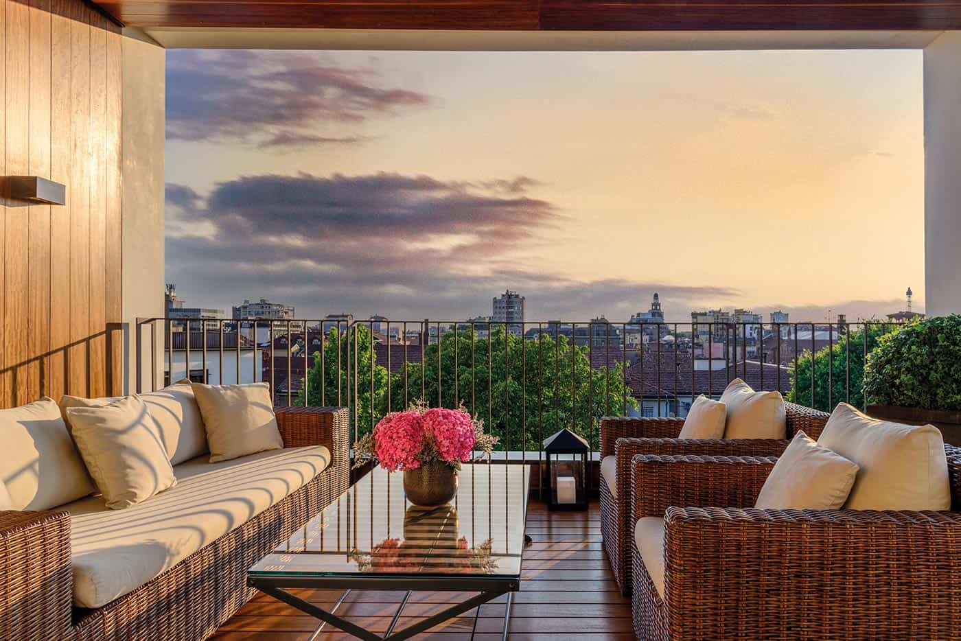 Rooftop terrace in Milan with cozy wicker furniture, pink flowers, and city skyline at sunset.