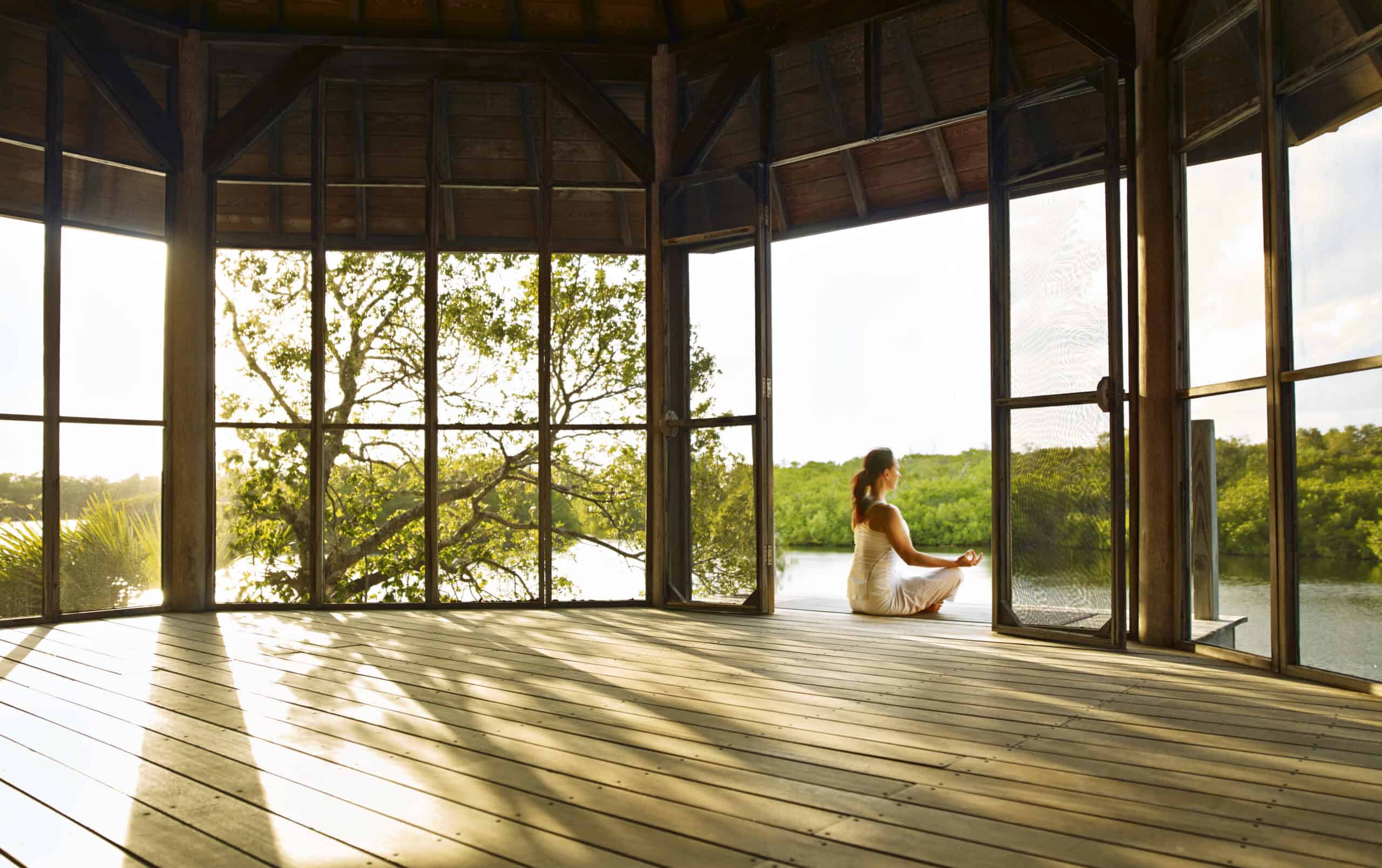 Relaxing woman meditating in a wooden pavilion overlooking a serene water landscape.