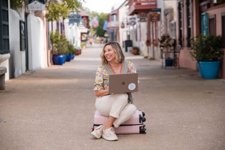 Woman sitting on suitcase with laptop in a vibrant, charming street, embodying travel and adventure.