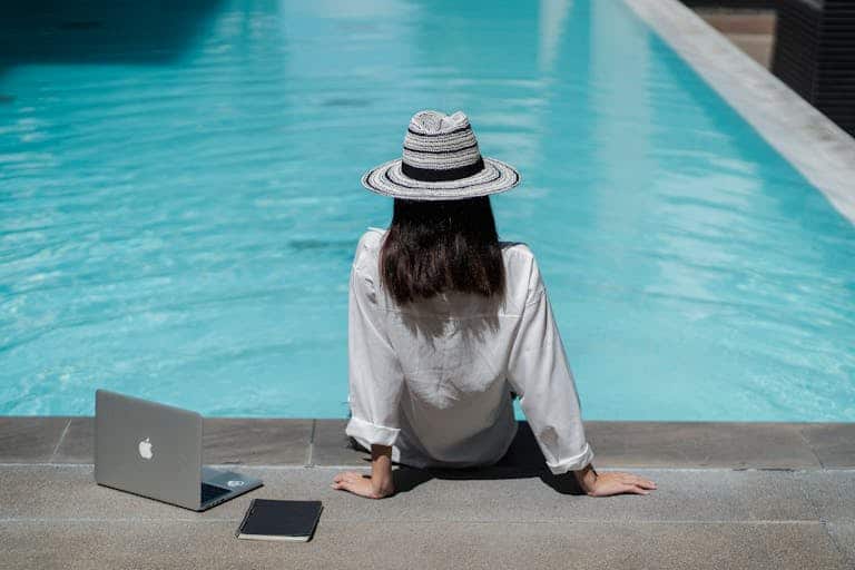 Woman sitting by a pool with a laptop and sun hat, enjoying leisure travel.